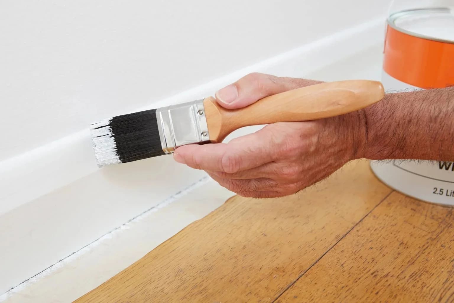 A decorator cutting in white gloss along a skirting board with a brush, paint tin on the floorboards alongside